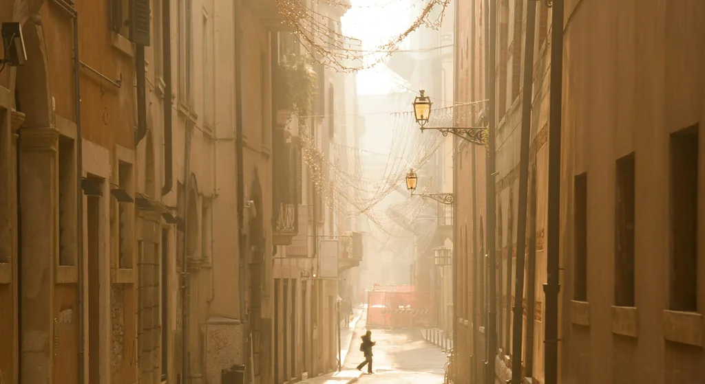 A misty European side street at sunrise, strung with lights overhead, with a single pedestrian walking past shuttered shopfronts